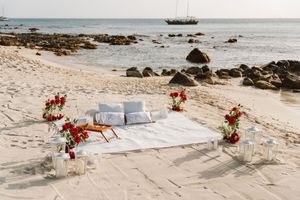 Romantic beach setup with white mattress, red roses, candles, and pillows by the ocean shore.