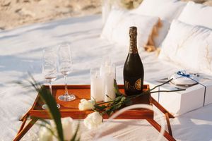 Romantic beach picnic setup with champagne bottle, two glasses, white candles, and roses on a wooden tray.