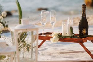 Elegant beach wedding setup with champagne flutes and a bottle on a wooden tray atop a white blanket by the sea.