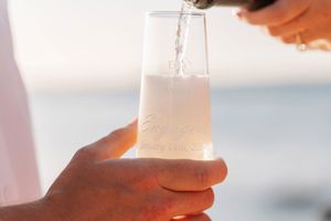Champagne being poured into an engraved stemless glass held by a hand near a serene ocean backdrop.