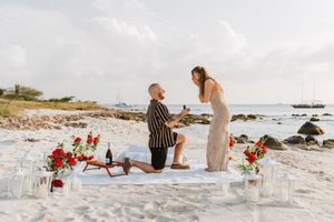 Man proposes to woman on sandy beach with red flowers and candles at sunset.