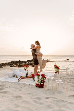 Couple sharing romantic beach sunset moment with red roses, candles, and champagne on white blanket by the ocean.