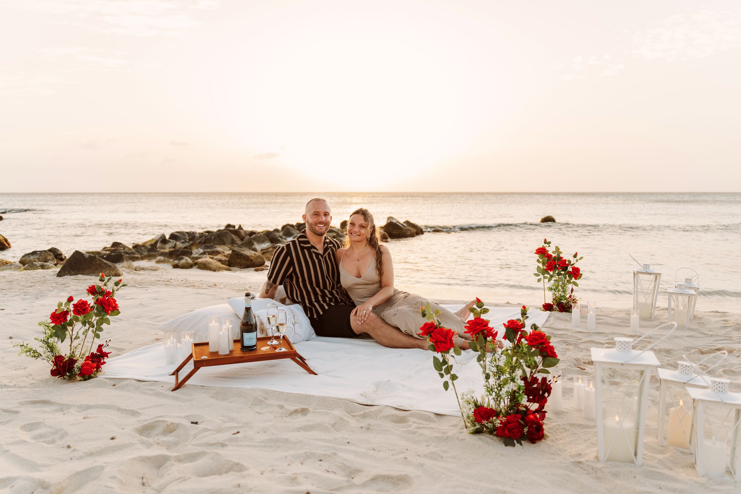 Couple sitting on white blanket with red roses and candles on sandy beach at sunset romantic proposal setup.