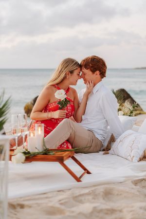 Romantic couple sharing an intimate moment on a beach picnic with candles, champagne, and a white rose at sunset.