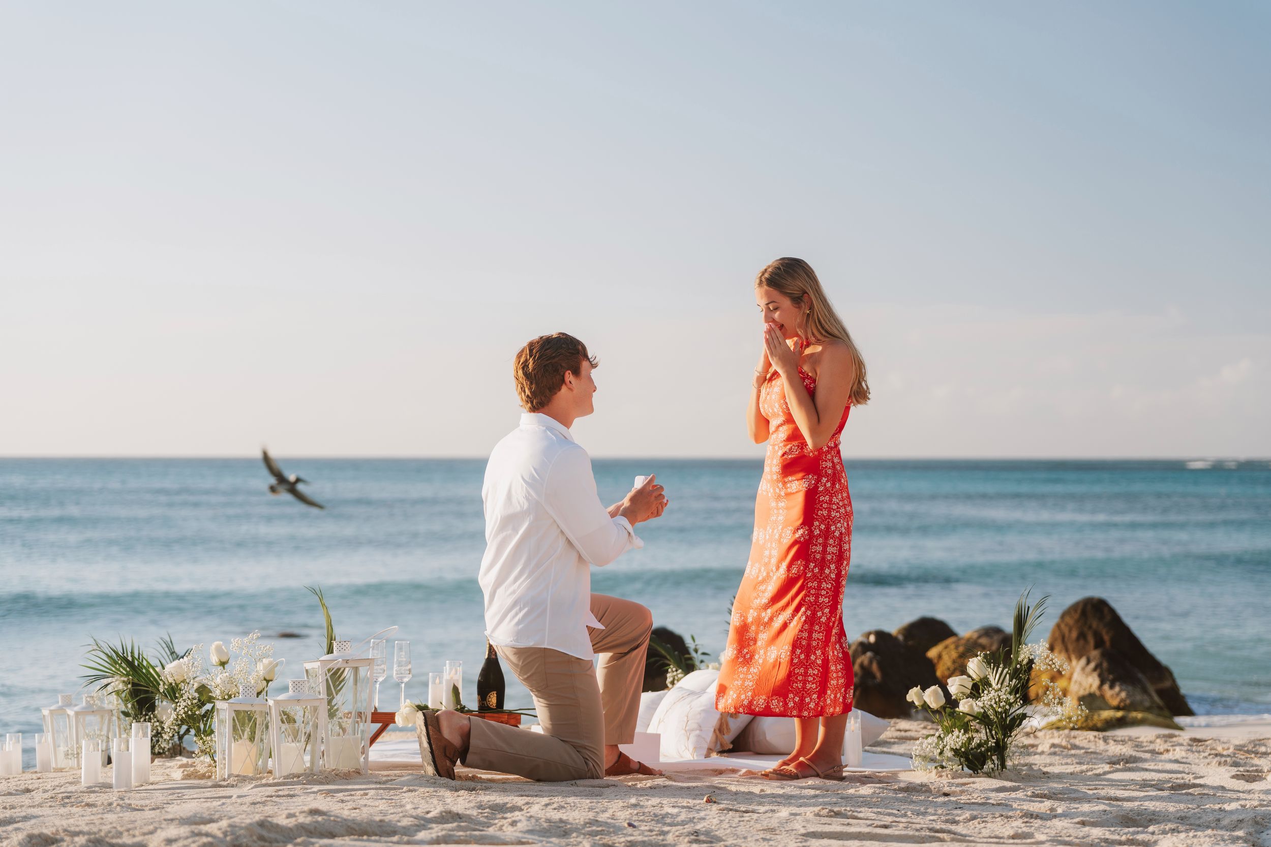 Man proposes to woman on a romantic beach setup with flowers and champagne at sunset by the ocean.