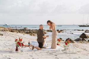 Man proposes on one knee to woman on sandy beach with red roses and champagne setup by the ocean.