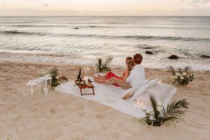 Romantic beach picnic at sunset with couple sitting on white blanket, candles, flowers, and wine by the ocean.