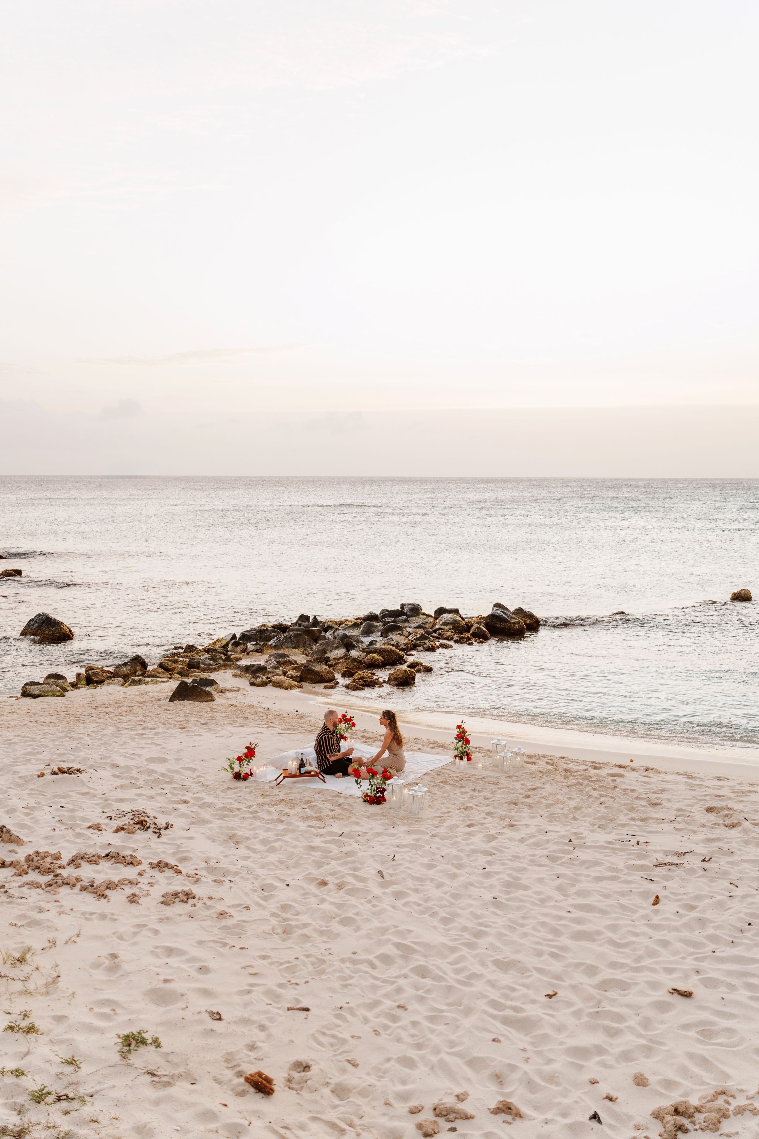 Couple sitting together on a white sandy beach at sunset, surrounded by floral arrangements near rocky shoreline.
