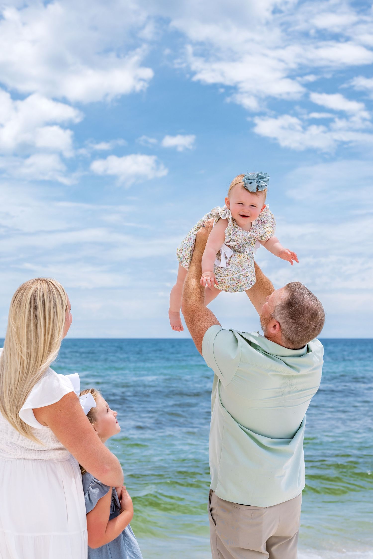 A father lifts his baby girl in the air on a sunny beach while the mother watches joyfully with the ocean behind them.