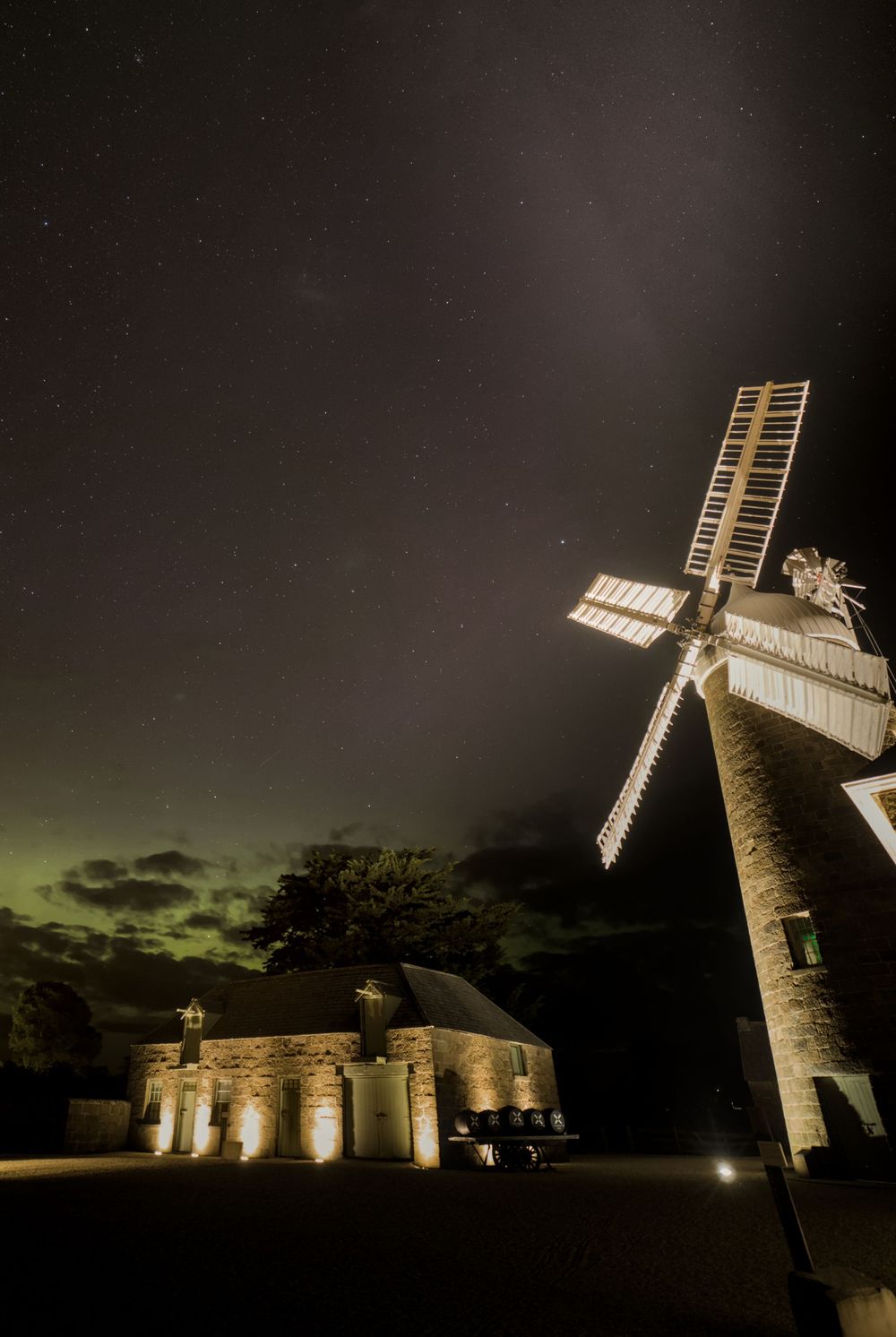 Historic windmill illuminated at night under starry sky with faint green aurora australis on horizon.