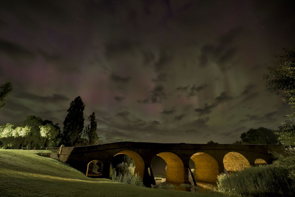 Historic stone arch bridge illuminated at night under a dramatic sky with colorful aurora borealis and clouds.