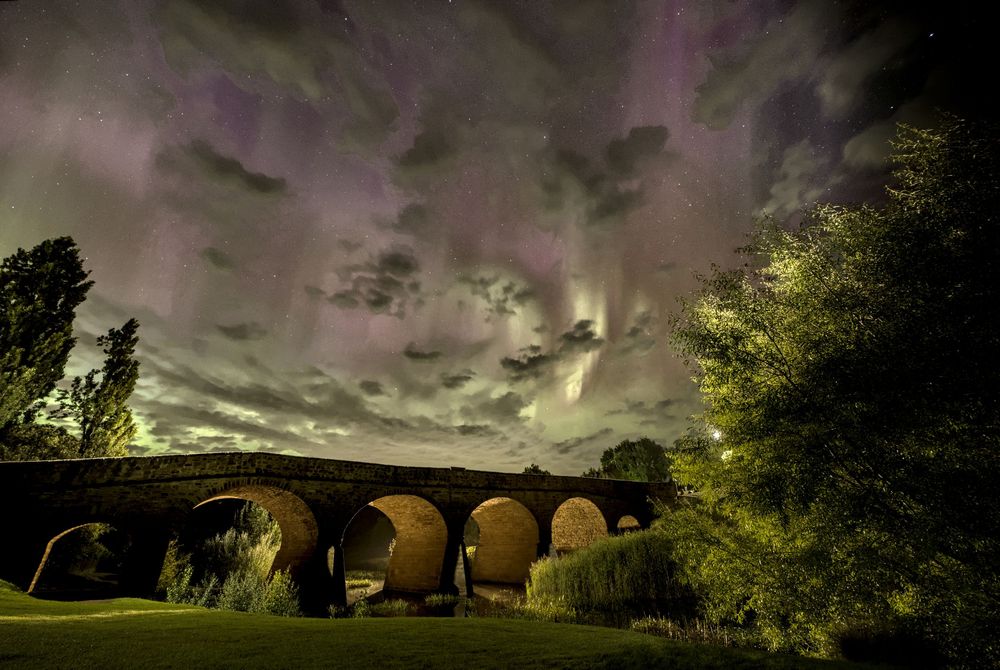 Magnificent purple aurora australis illuminating cloudy night sky above historic multi-arch stone bridge with trees.