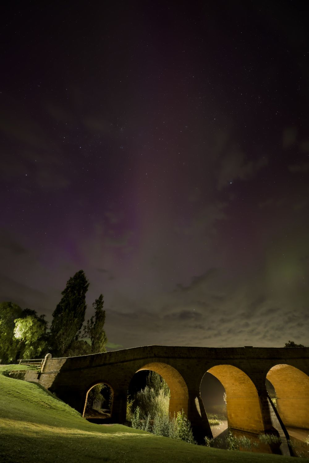 Historic stone arch bridge illuminated at night with vibrant aurora borealis and stars in the dramatic purple sky.