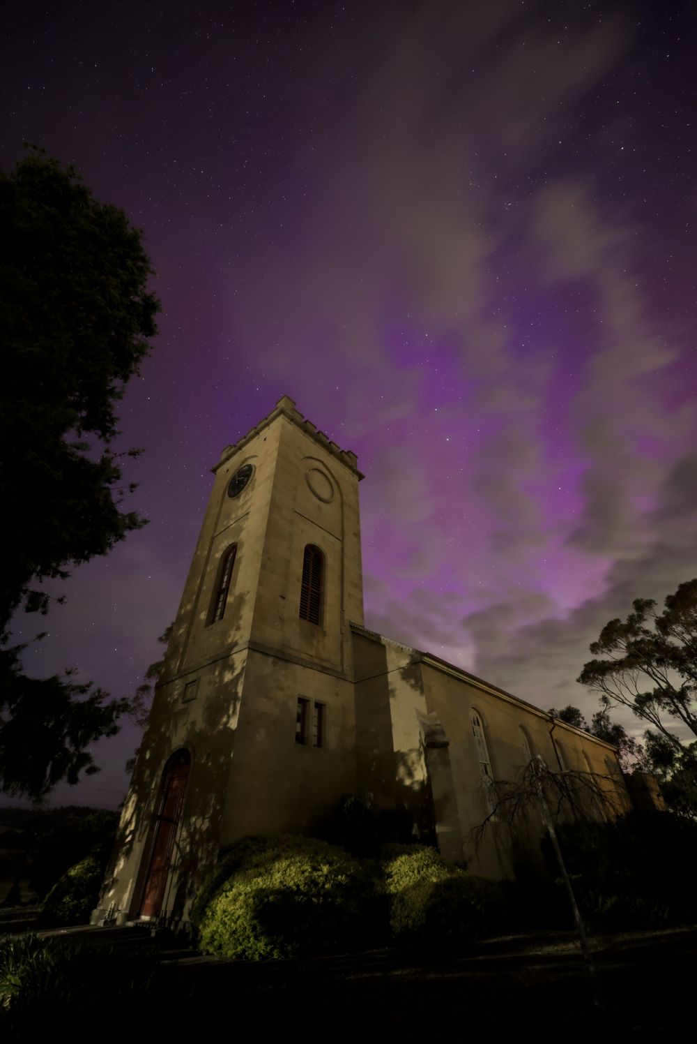Historic stone church tower under a dramatic purple aurora borealis night sky with glowing clouds and stars.