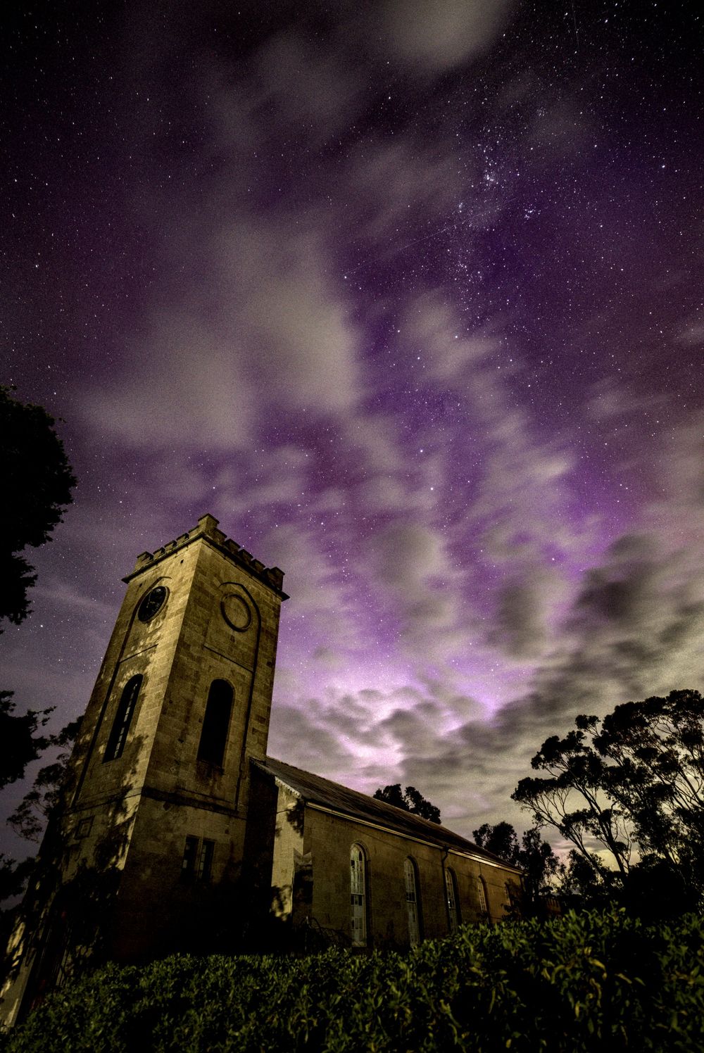 Historic stone church tower under dramatic purple aurora australis and starry night sky.