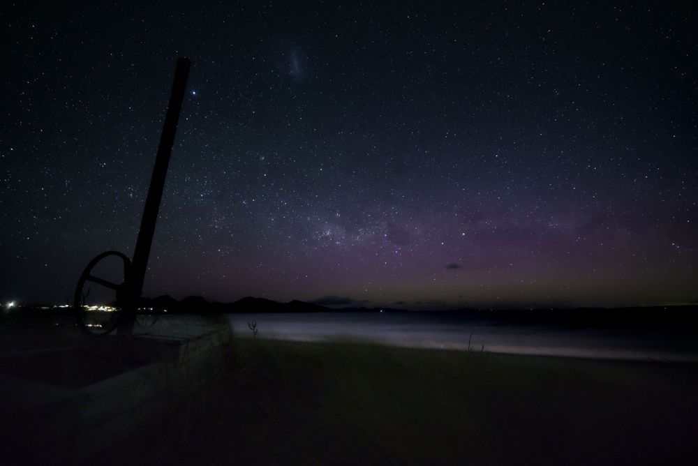 Milky Way and aurora australis over dark beach with silhouetted post, starry night sky.