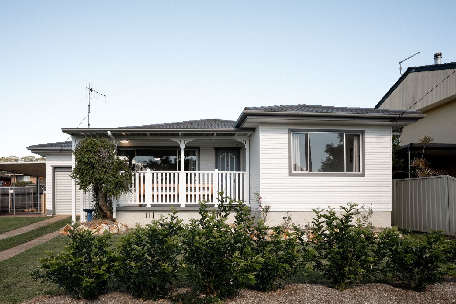 White Australian suburban home with front porch, white picket fence, and lush green garden shrubs under clear blue sky.