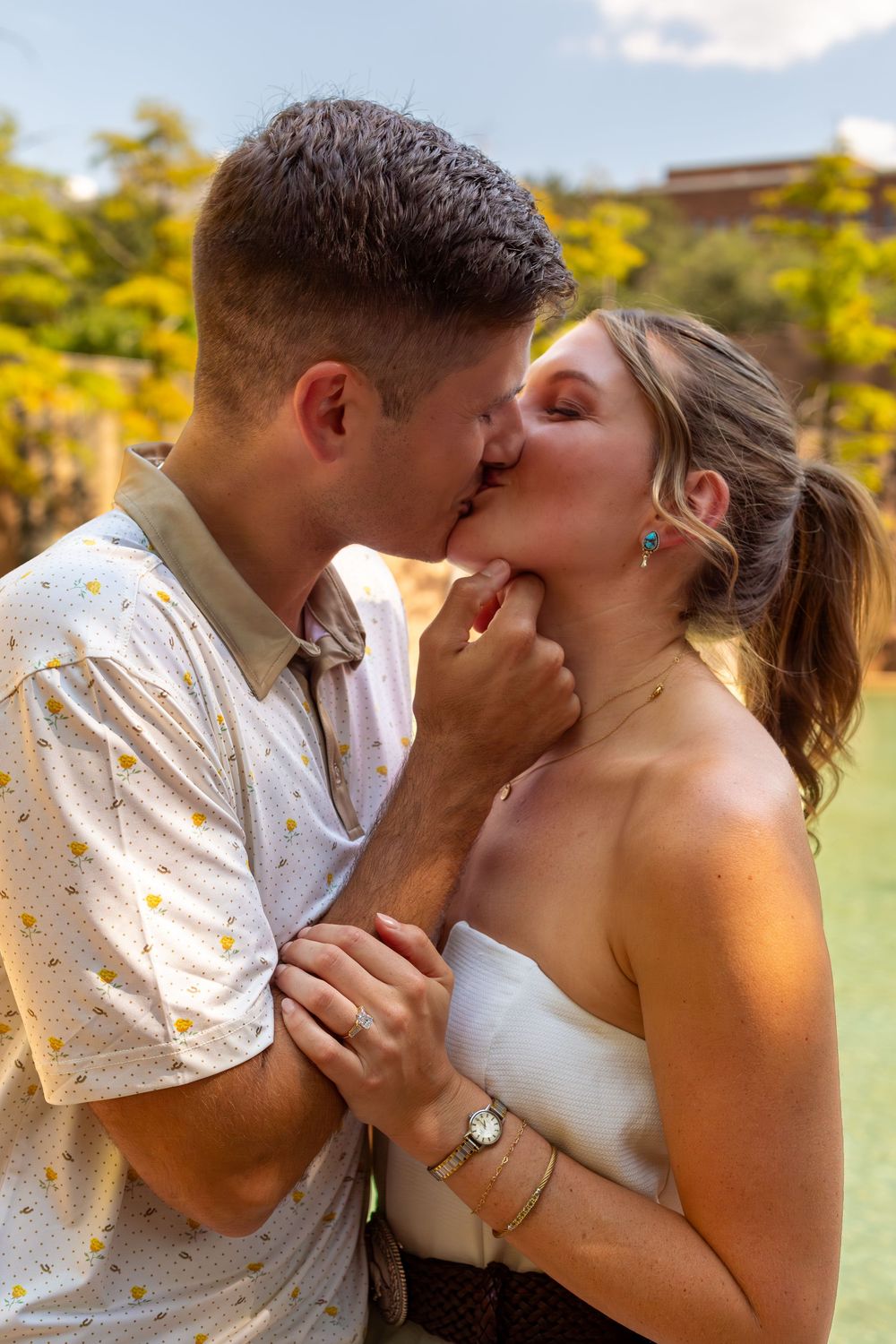 Couple sharing a romantic kiss outdoors, woman in white dress with gold jewelry, man in floral shirt, lush green background.