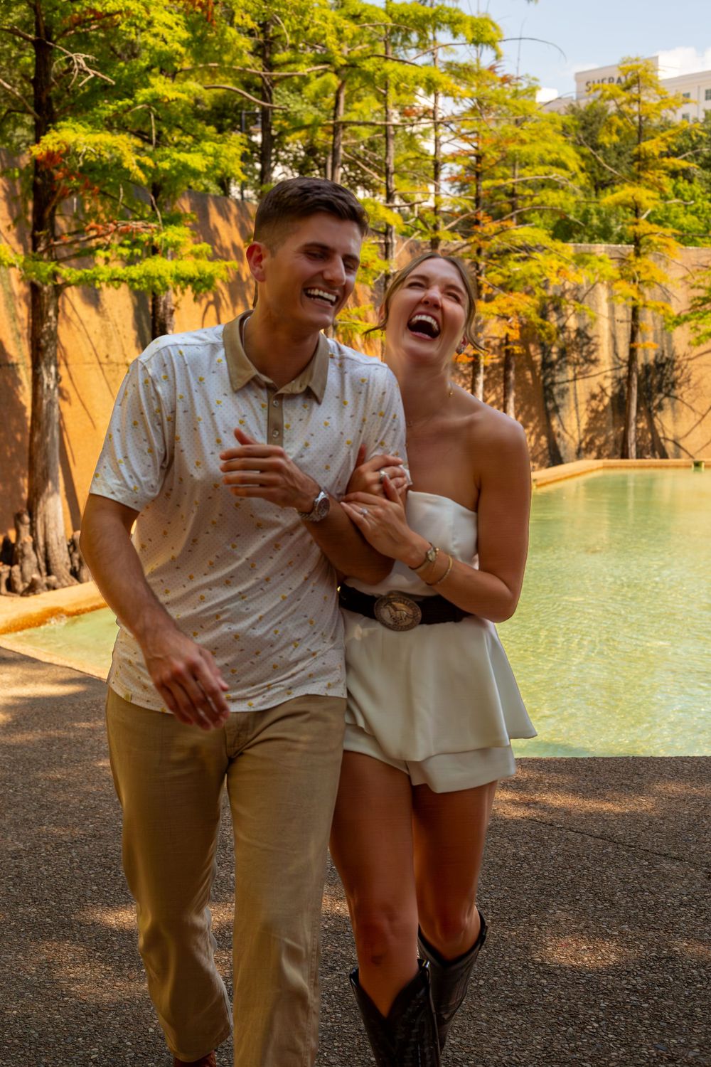 Happy couple laughing and walking together near a scenic pool with autumn trees.