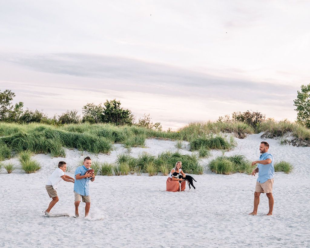 Family playing on white sand beach at sunset with green dunes, two men tossing ball while others relax nearby.