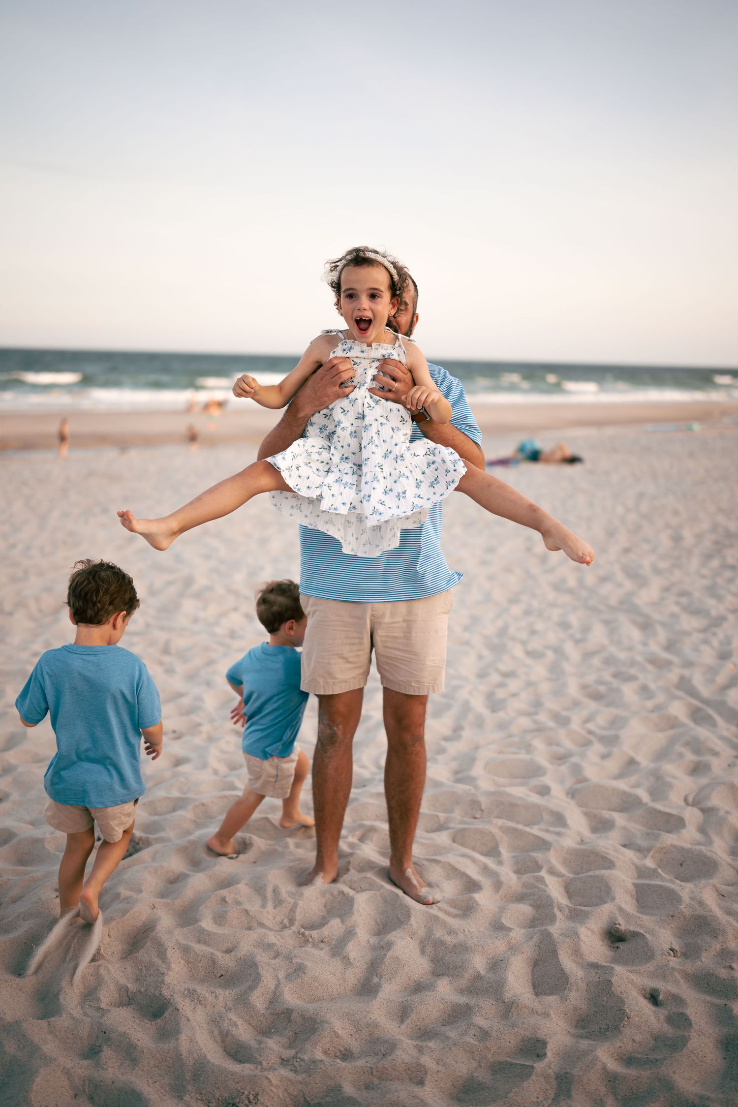 Father lifting excited girl on beach while two boys play in sand at sunset.