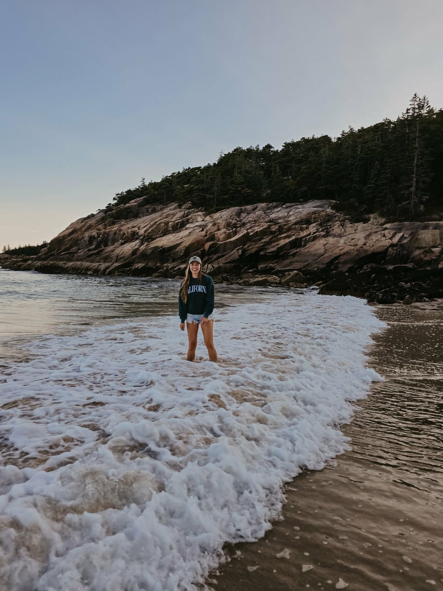 Woman standing in ocean waves on a sandy beach with rocky cliffs and pine trees.
