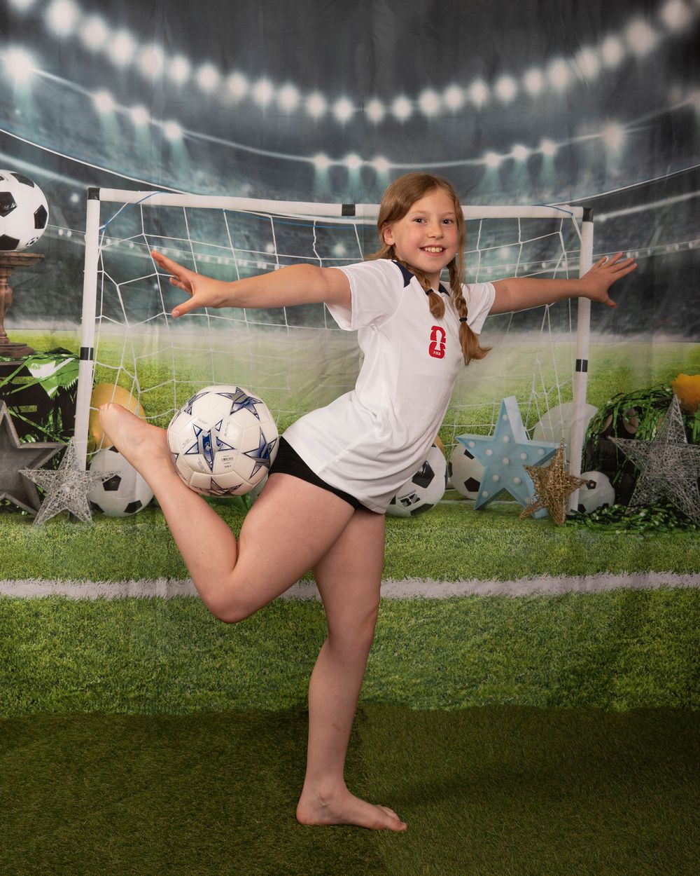 Young girl in white soccer jersey poses with soccer ball in front of stadium backdrop with goal net.