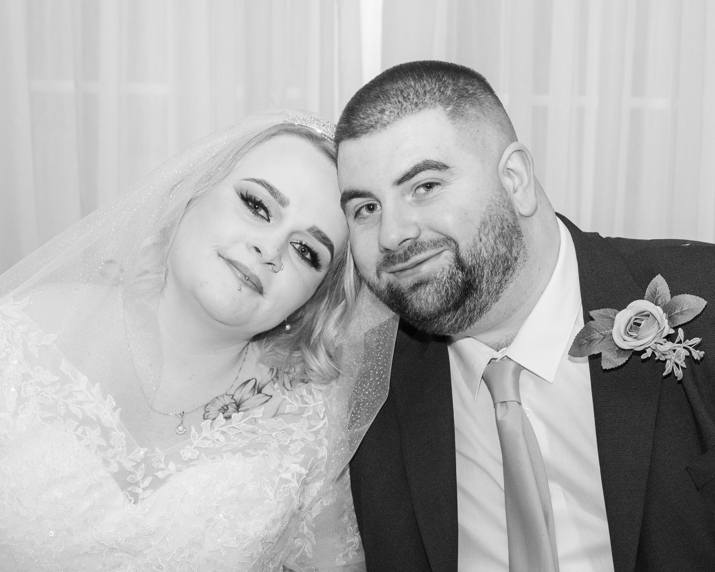 Black and white wedding photo of smiling bride in lace gown and groom in suit with boutonniere posing cheek to cheek.