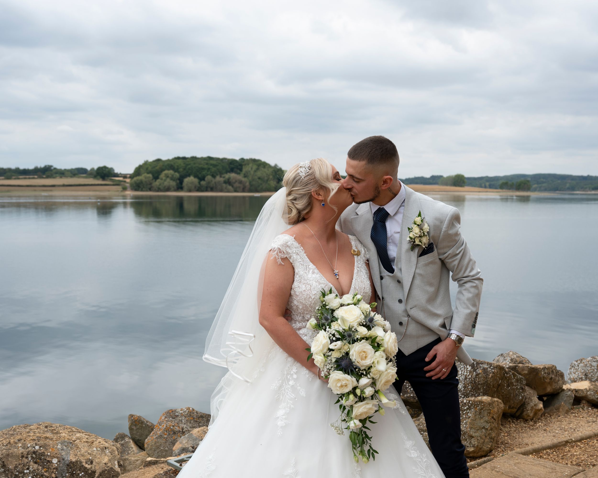 A romantic wedding couple embraces near a serene lakeside setting with cloudy skies and natural scenery in the background.