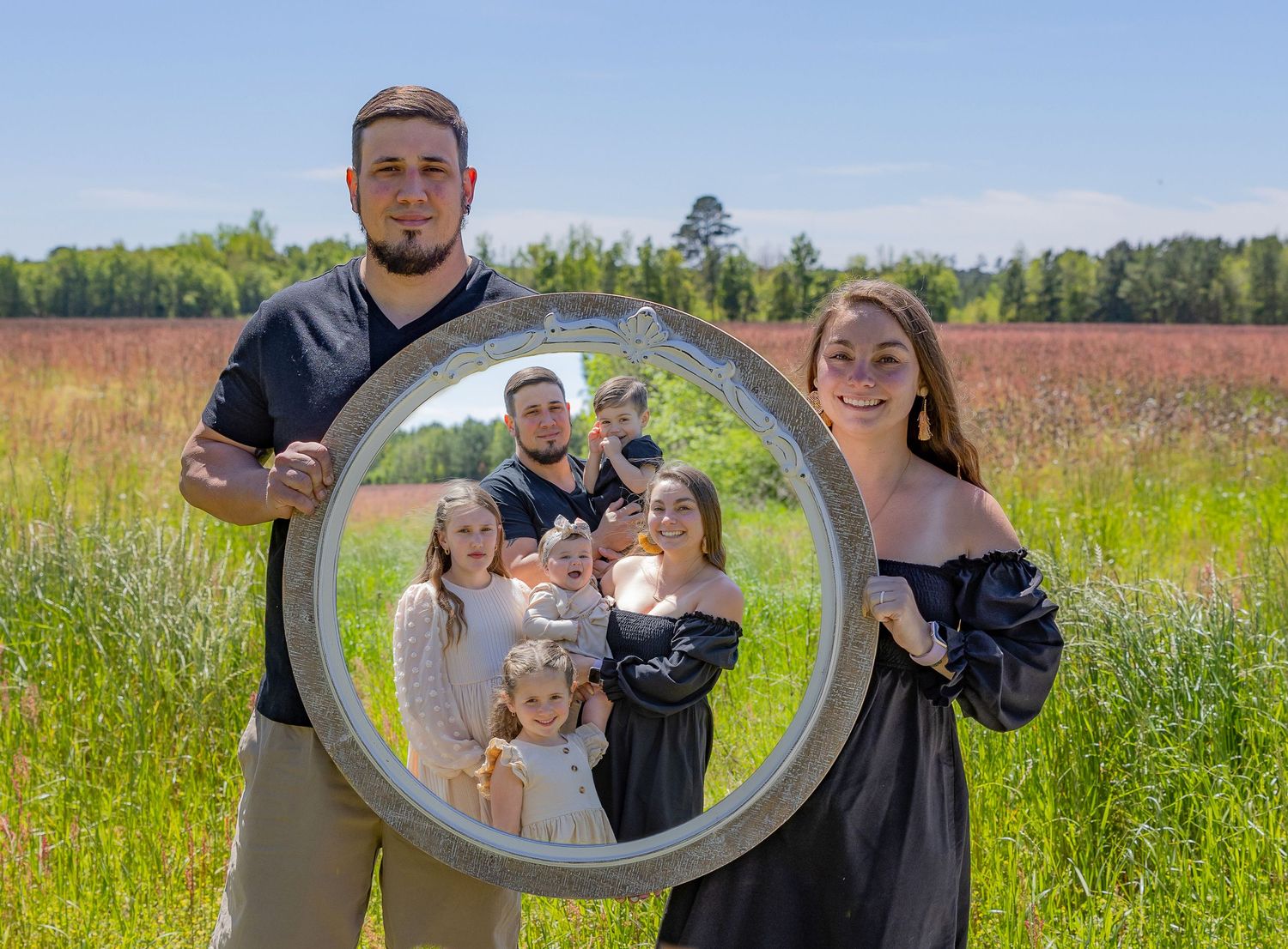 Couple poses playfully with a circular metal frame in a sunny meadow field.