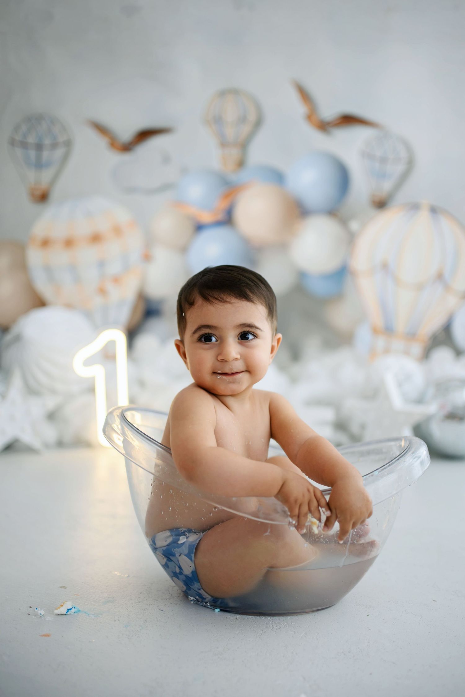 Smiling baby sits in a clear tub at a first birthday cake smash photoshoot with hot air balloons, blue balloons and a glowing number one.