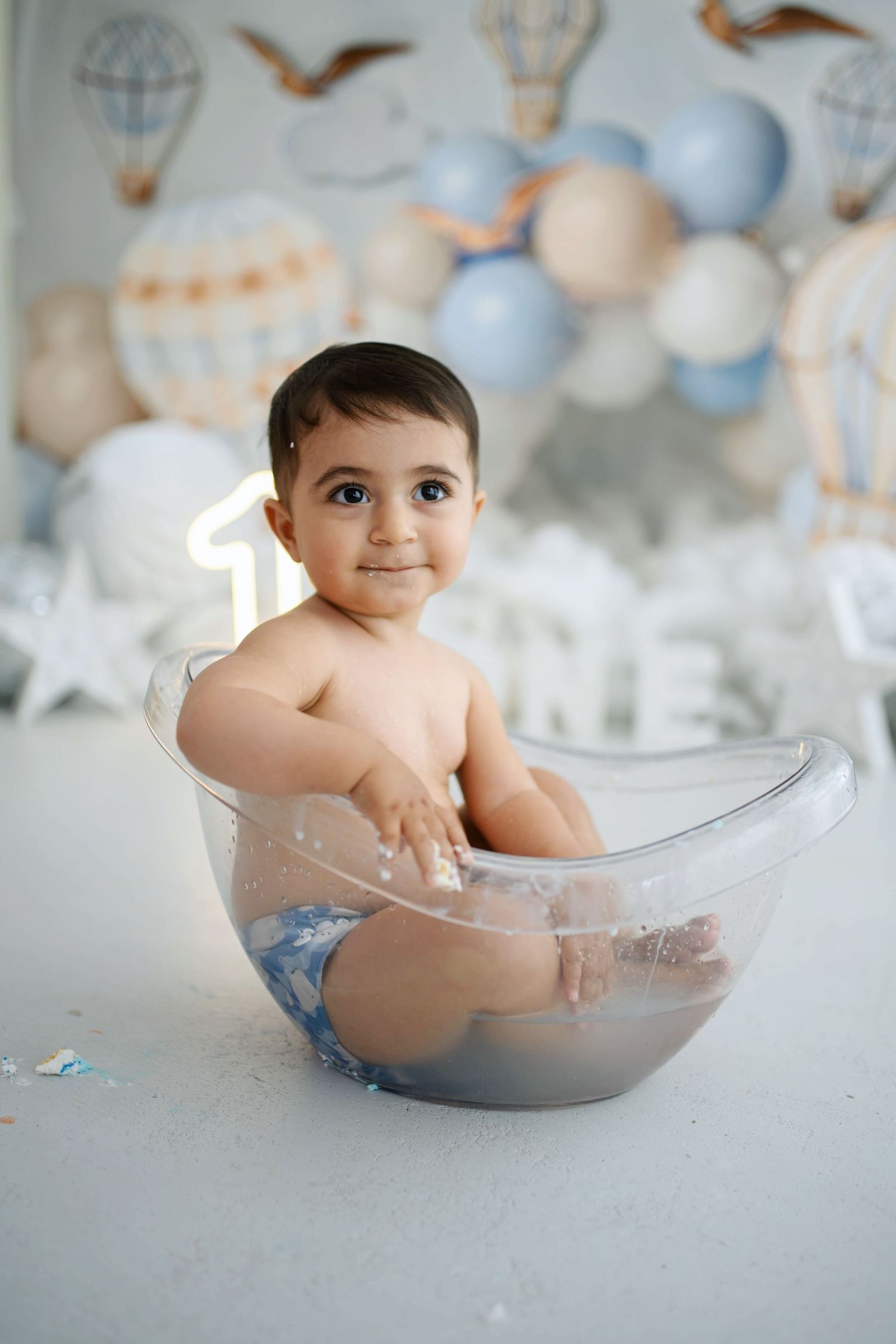Smiling toddler sits in a clear bathtub during a first birthday cake smash photoshoot with hot air balloon decor and a glowing number one.
