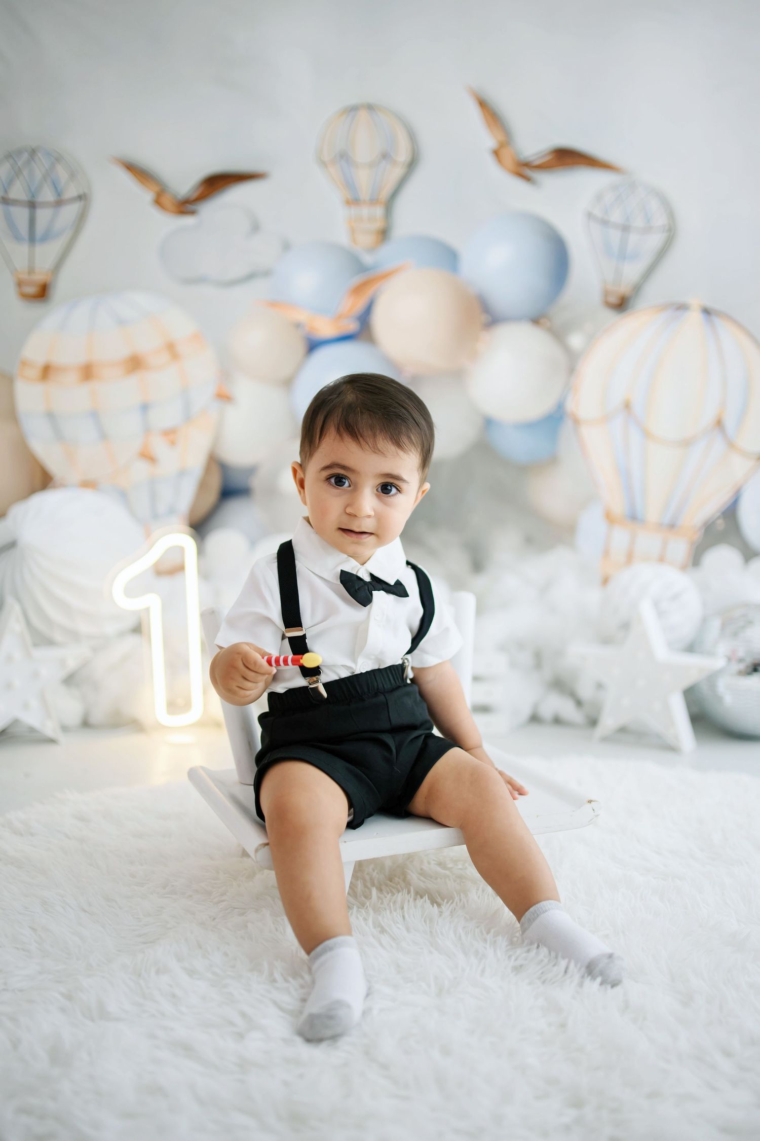 Toddler boy in a white shirt, bow tie and black suspender shorts sits on a white stool at a hot air balloon first birthday photoshoot.