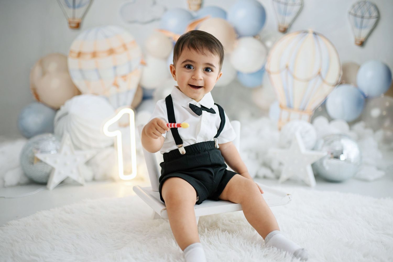 Smiling baby boy in suspenders and bow tie sits on a white fluffy rug beside a glowing number one for a hot air balloon first birthday.