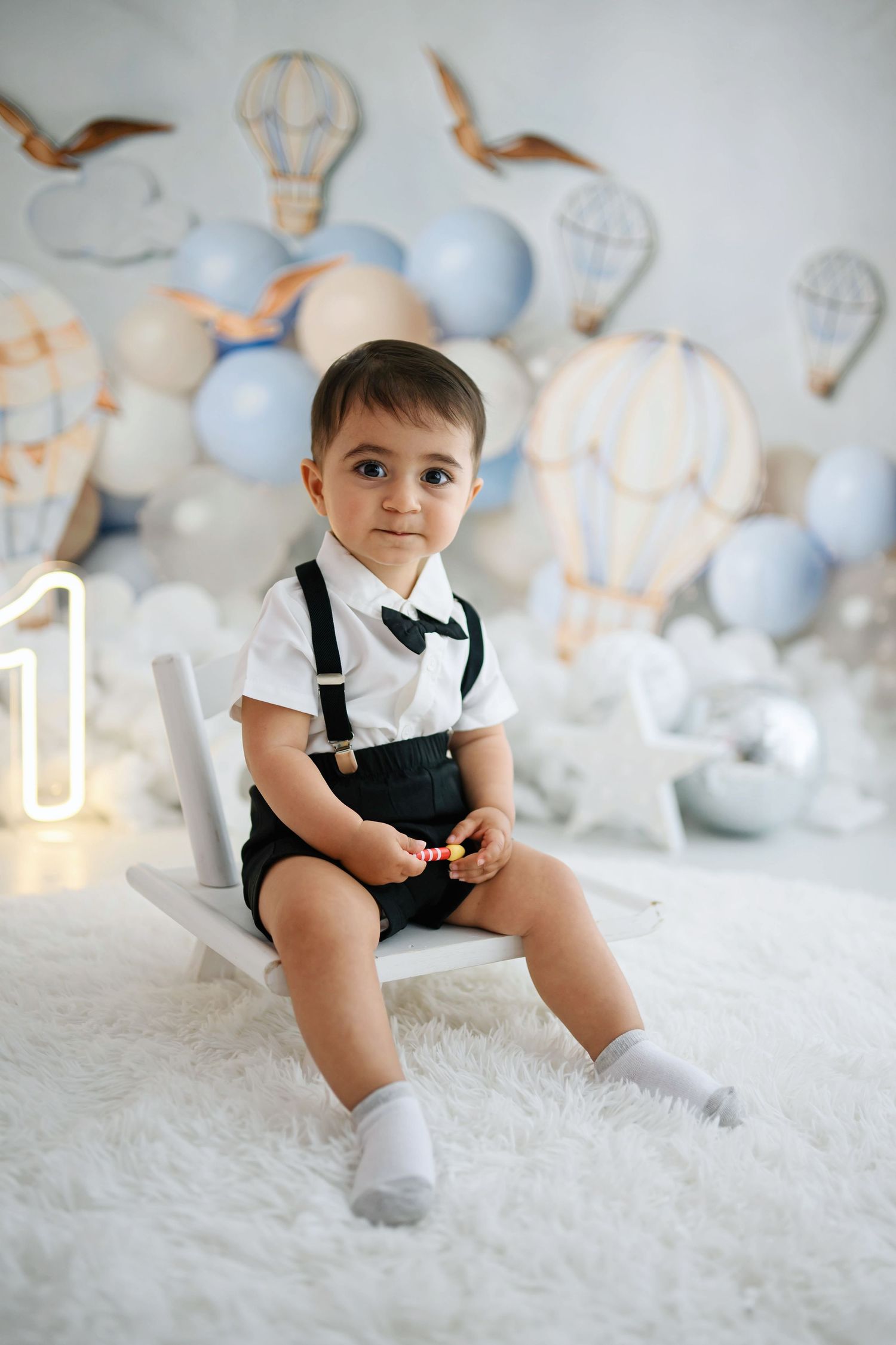 Toddler in white shirt, bow tie and black suspender shorts sits on a small white chair at a hot air balloon first birthday photoshoot.