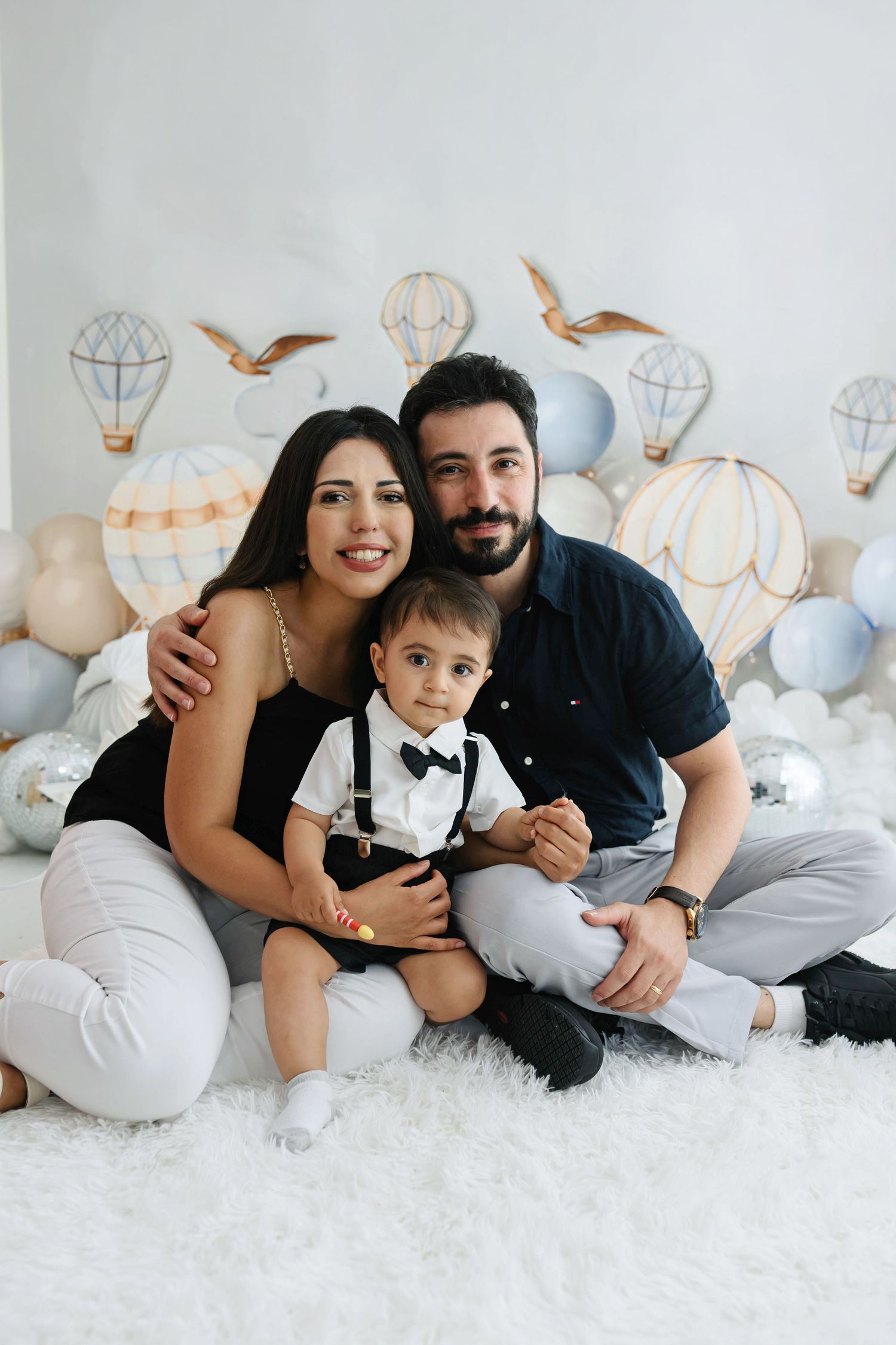Smiling couple sits with their toddler in a bow tie and suspenders on a fluffy white rug beside balloons and hot air balloon decor.