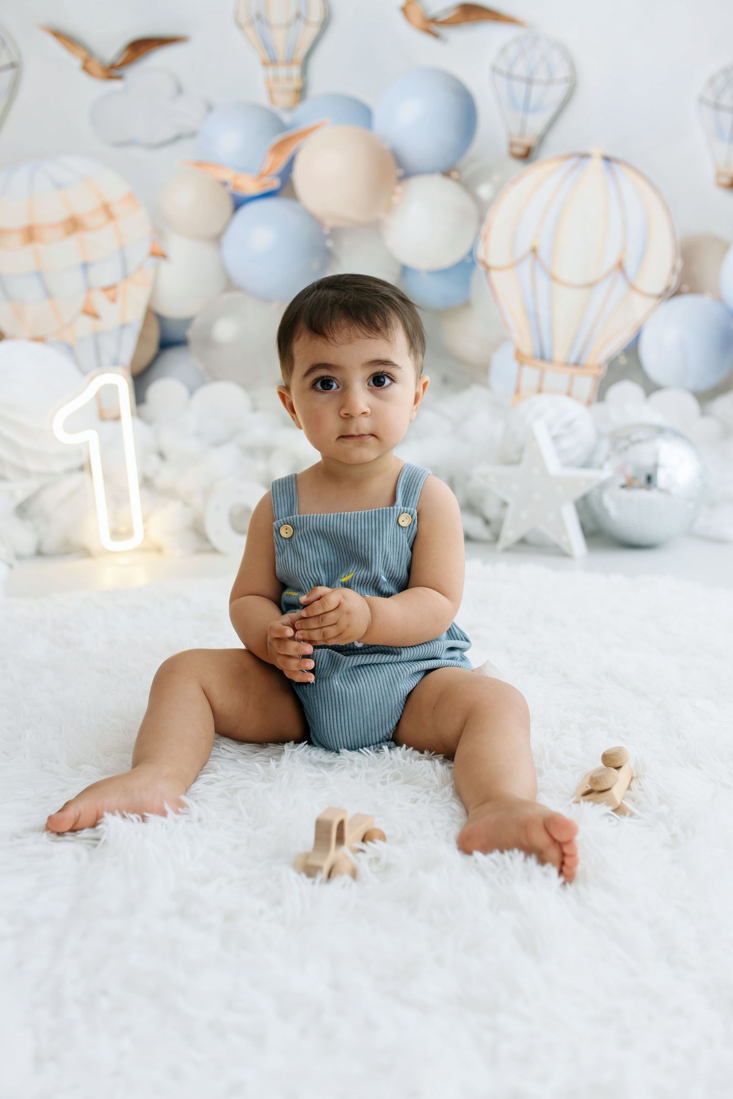 Baby in blue denim overalls sits on a fluffy white rug at a first birthday cake smash photoshoot with hot air balloons and balloons.