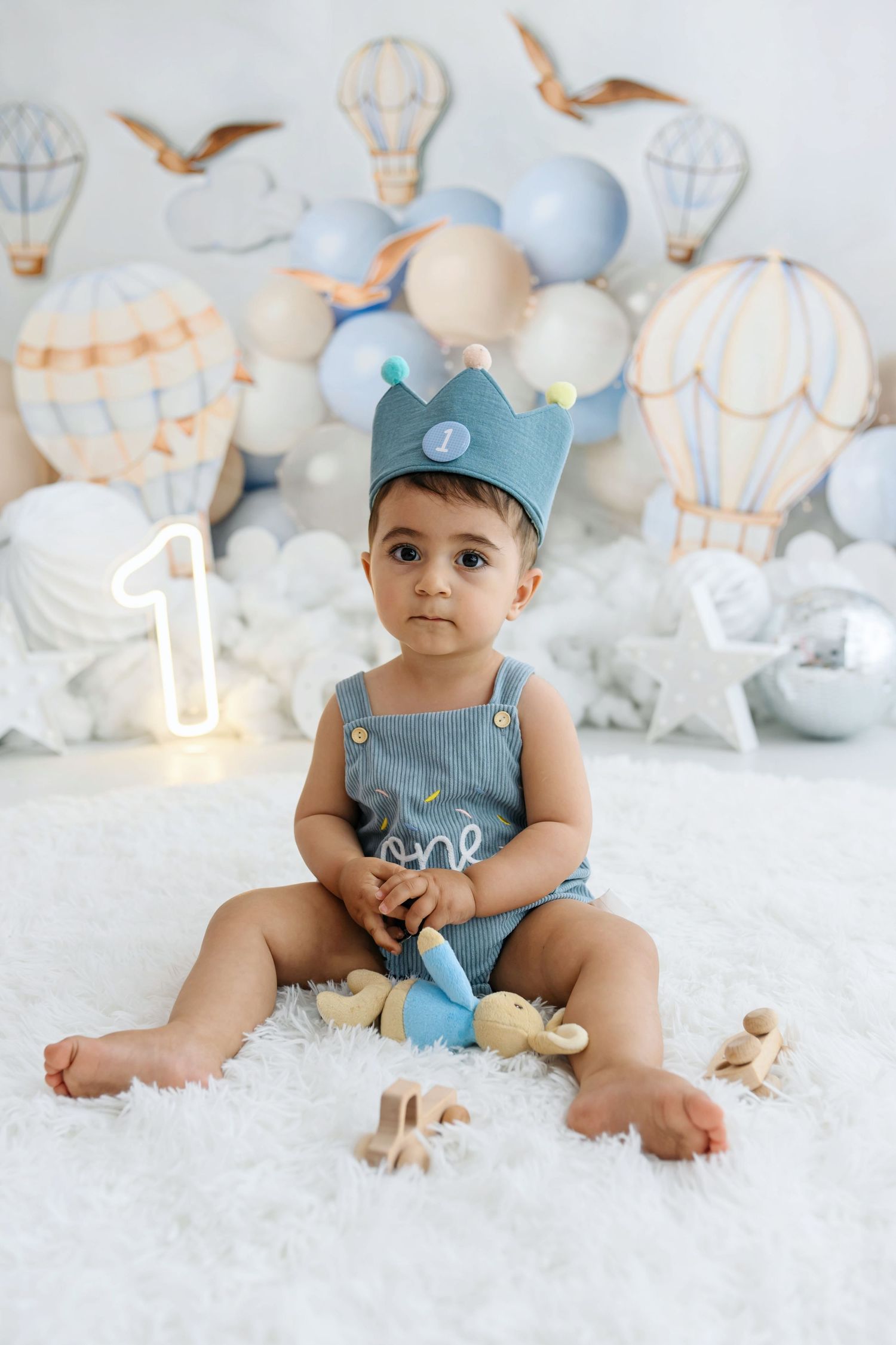 Baby in a blue crown and denim overalls sits on a fluffy white rug at a first birthday cake smash set with hot air balloons.
