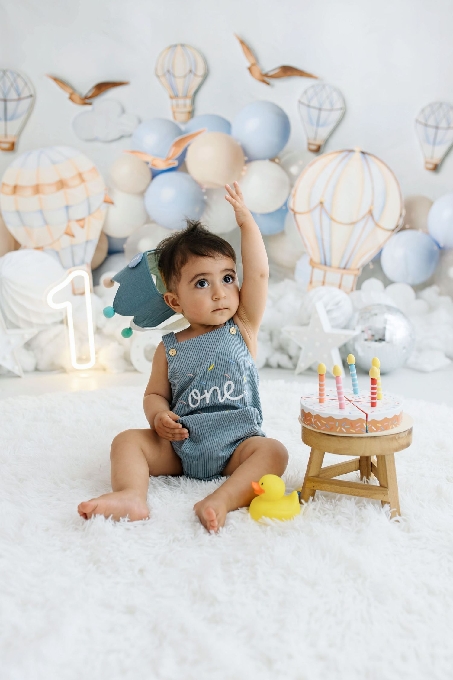 Baby in a gray romper that says one sits on a white fluffy rug beside a birthday cake, rubber duck and hot air balloon decor.