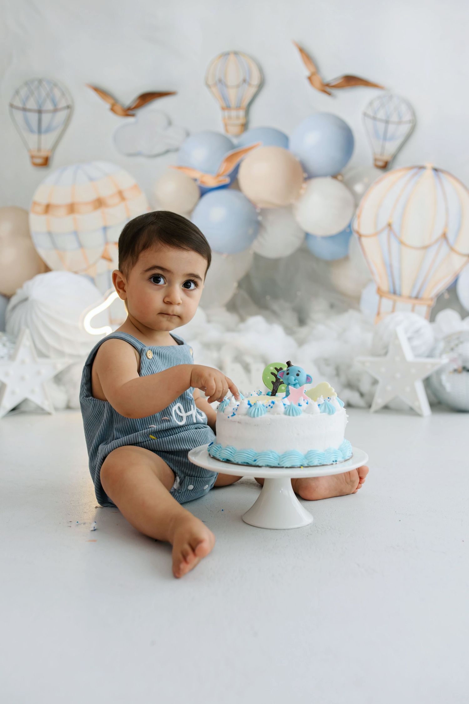 Baby in a blue romper sits beside a small blue cake at a cake smash photoshoot with hot air balloon decor and clouds.