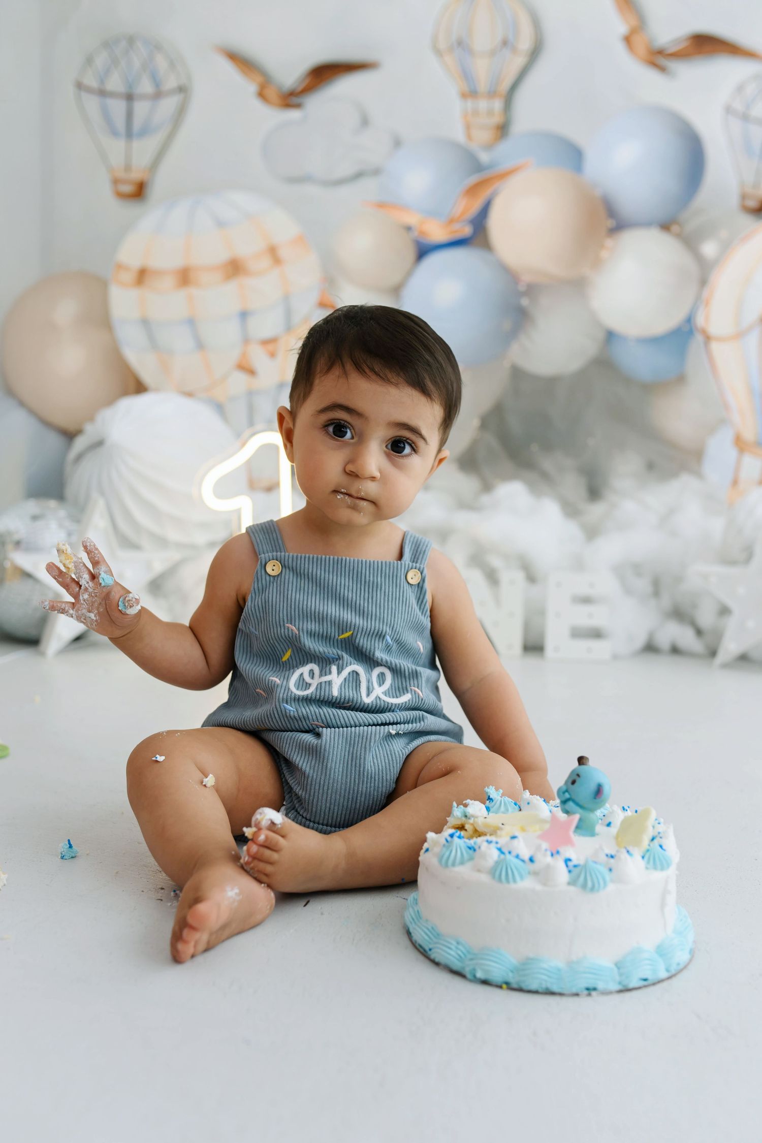 Baby in a denim overall with "one" on it sits beside a blue smash cake at a hot air balloon first birthday photoshoot.