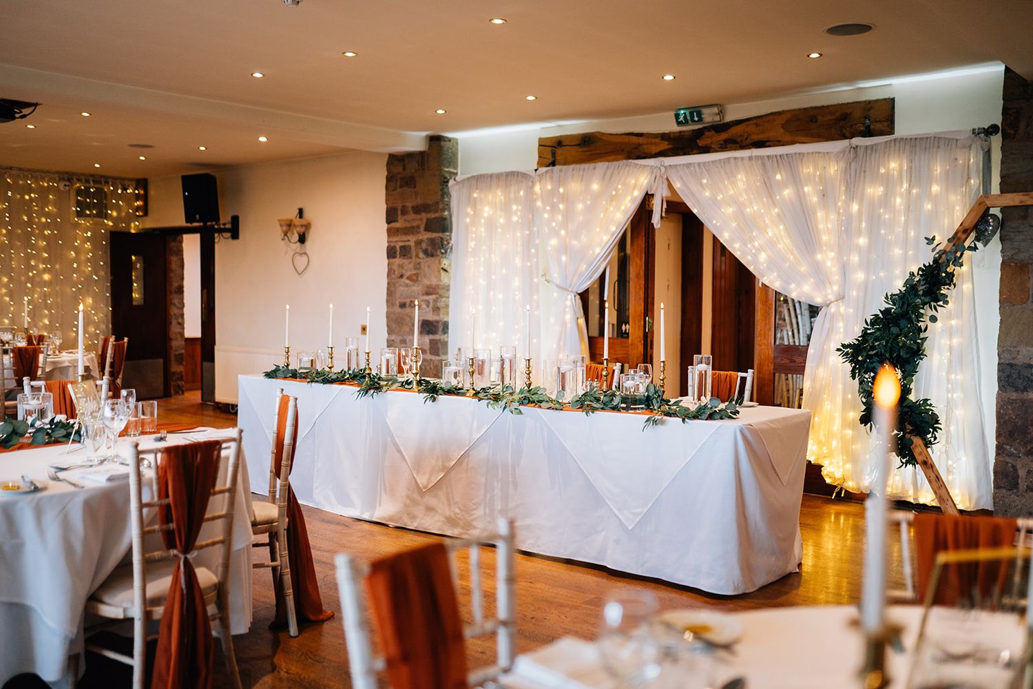 Rustic wedding reception venue with a long white-draped head table, greenery garland, candles and warm fairy light curtains.