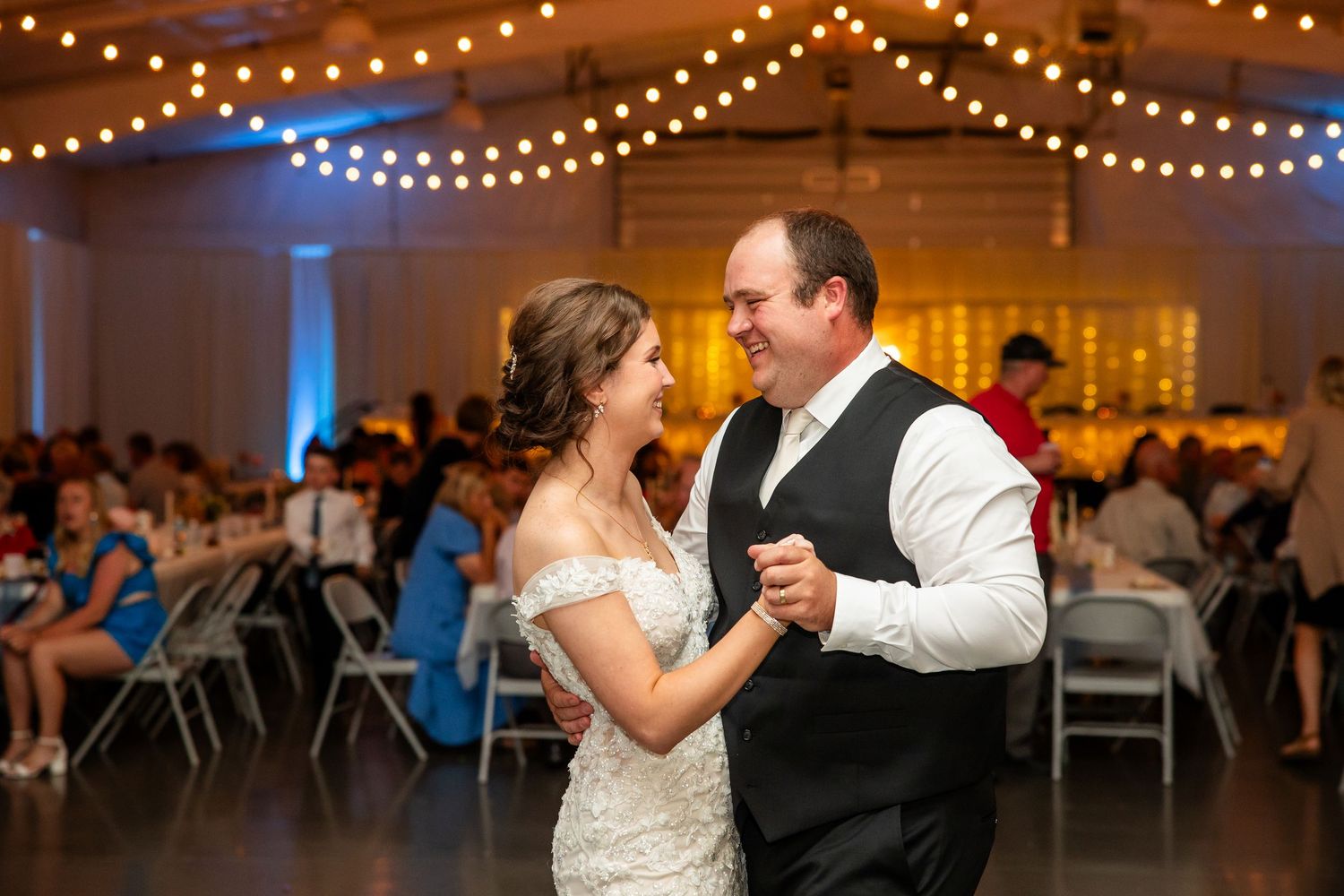 Bride in a white lace wedding dress and groom in a black vest share a first dance under string lights at a wedding reception.