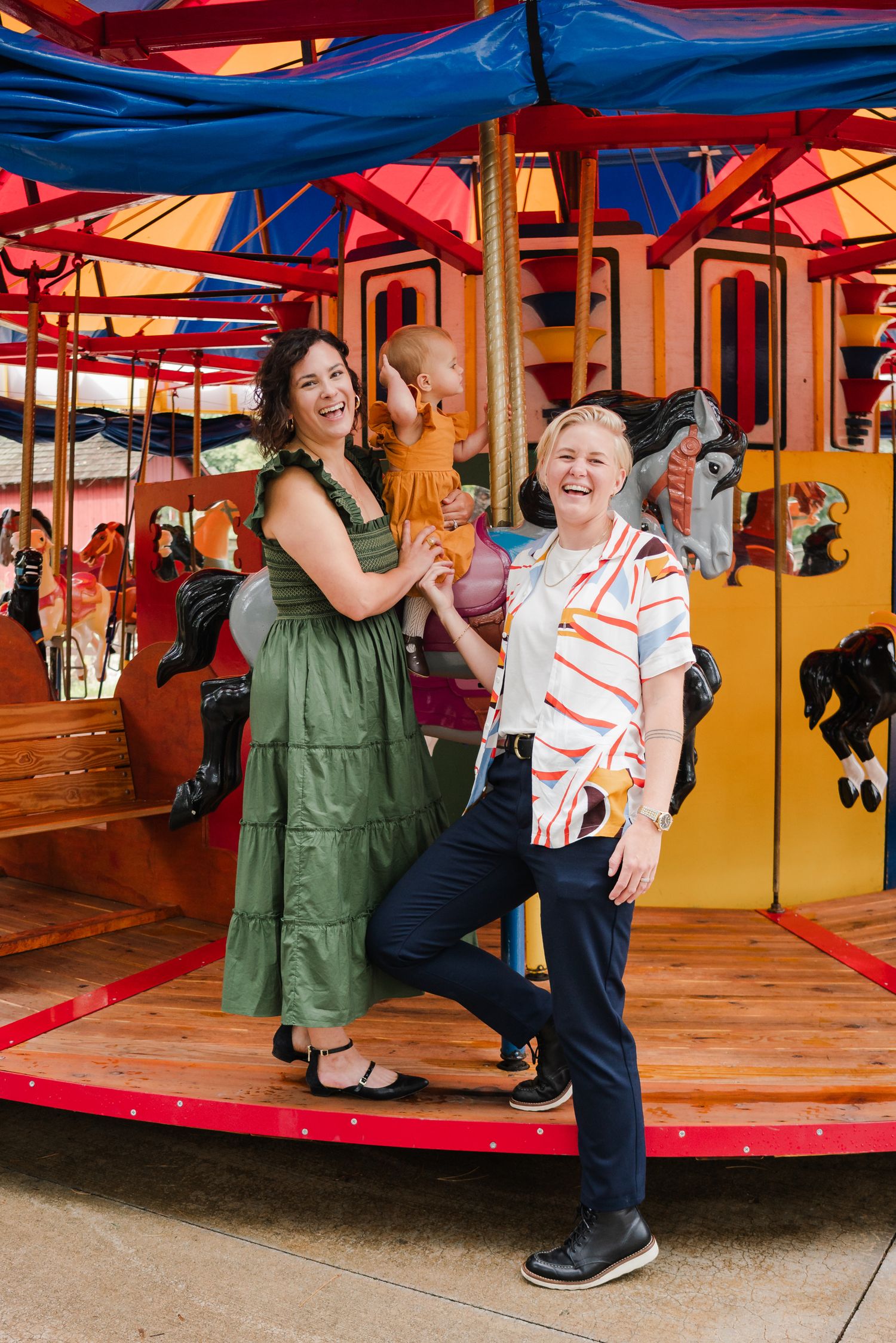 Two smiling women and a baby pose together on a colorful carousel at an amusement park during a joyful family photo session.