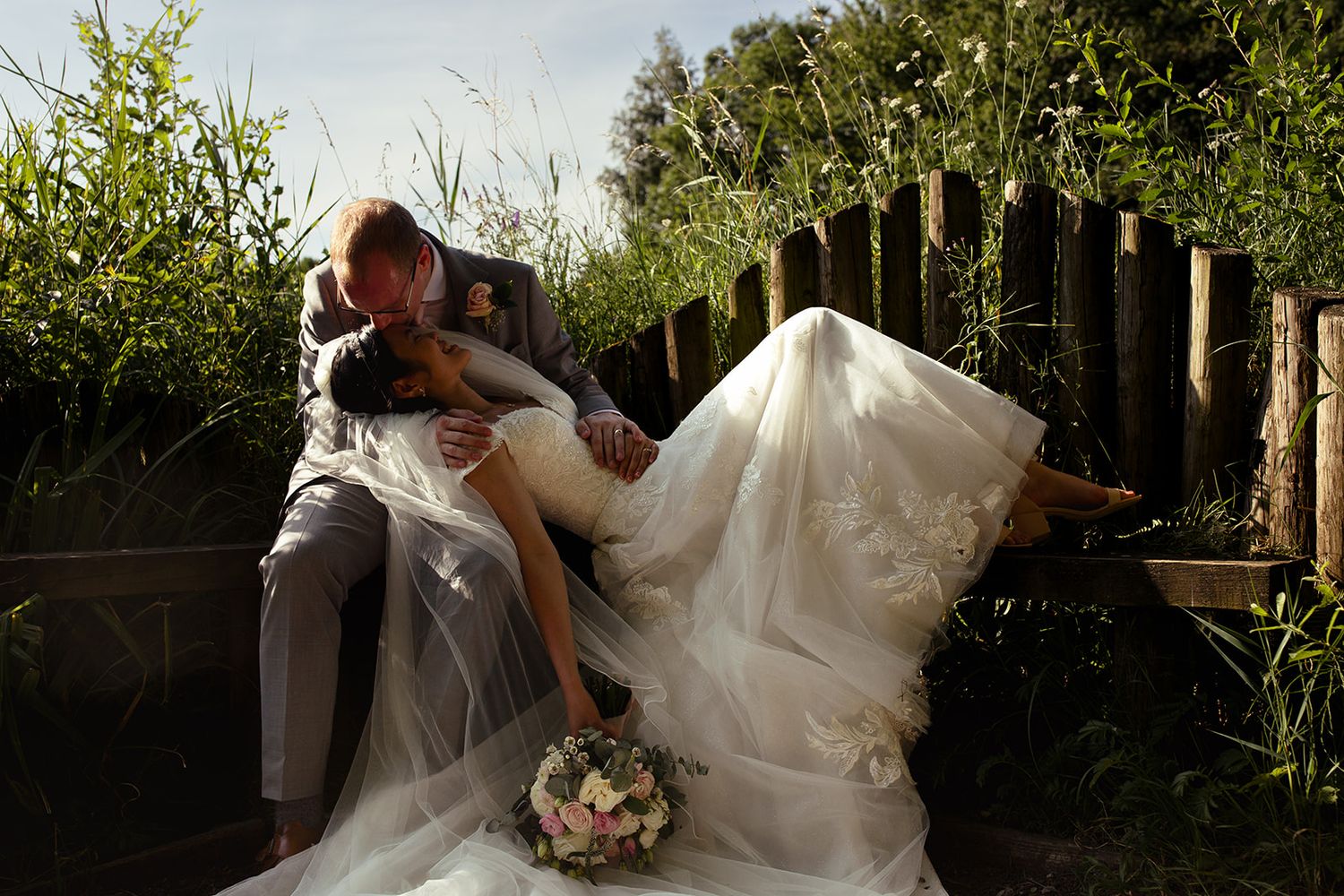 Bride in a flowing white wedding gown reclines in her groom's arms on a wooden bench, holding a pastel bouquet amid lush greenery.