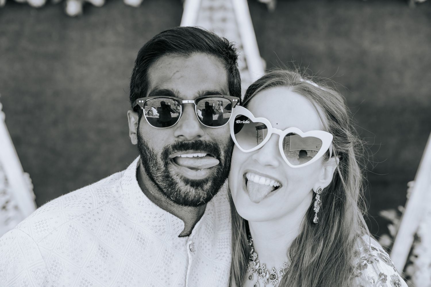 Black and white photo of a smiling couple in sunglasses, the woman in heart-shaped glasses sticking out her tongue, posing cheek to cheek.