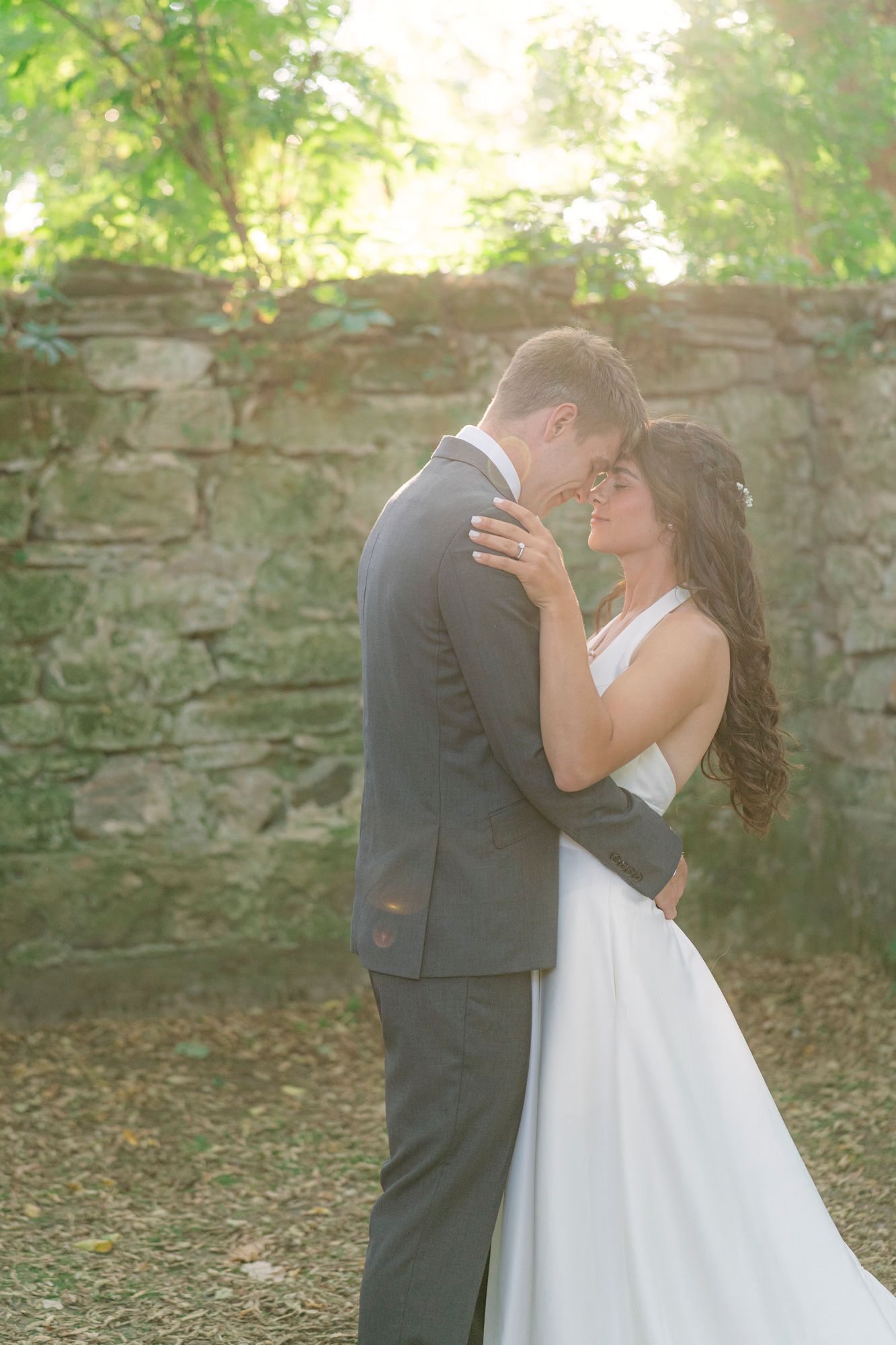 Bride in a white gown and groom in a gray suit embrace foreheads together in front of a rustic stone wall at a sunlit outdoor wedding.