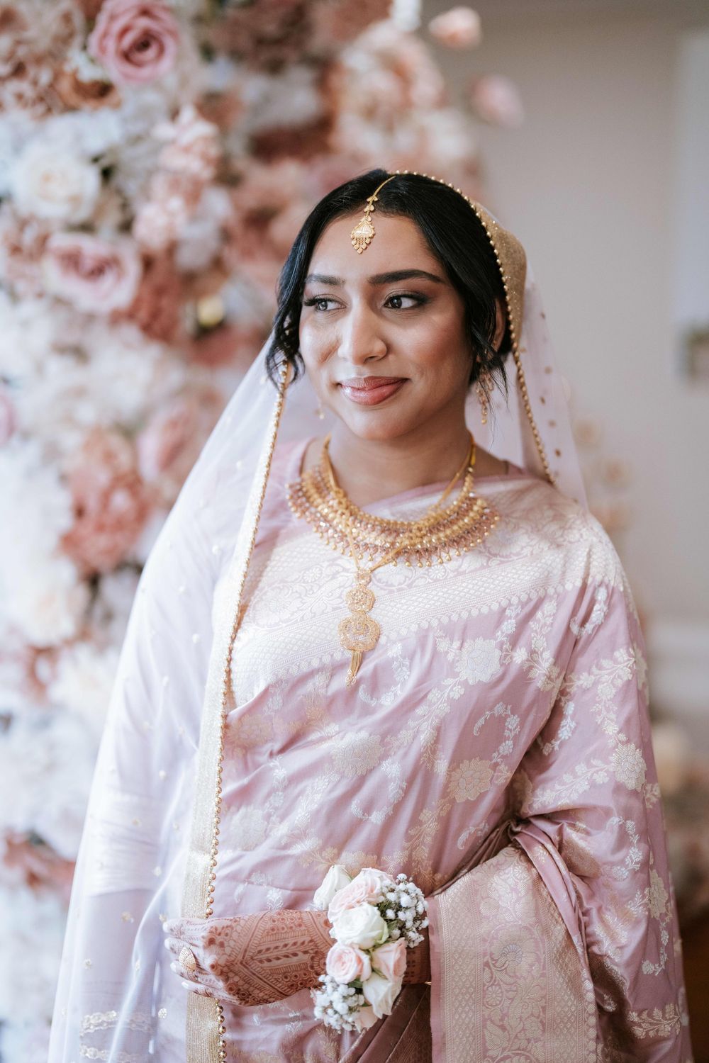 Bride in blush pink saree and veil in front of a floral wall before her Nikah ceremony