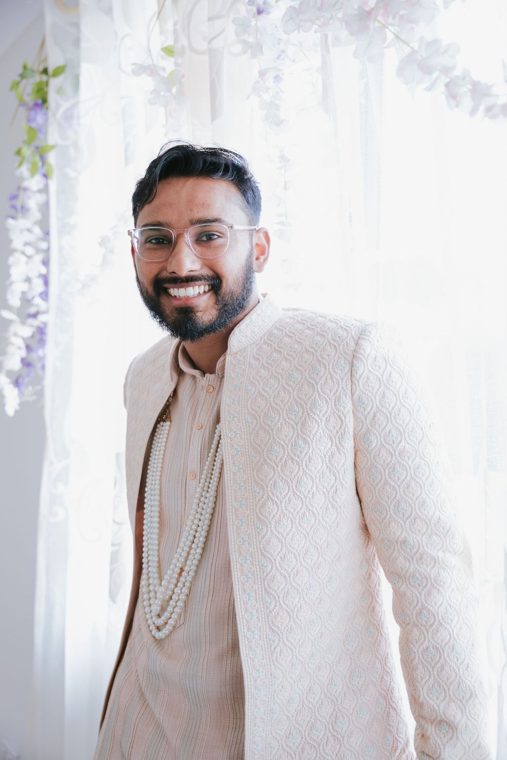 Smiling bearded man in glasses wearing a cream embroidered jacket and layered pearl necklace, standing before sheer curtains and white flowe