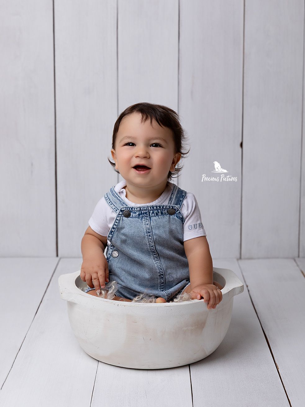 Joyful baby boy in denim dungarees sitting in a white rustic bowl, professional sitter milestone photography at Precious Pictures